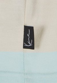 Chemise à manches courtes en coton crème et bleu clair avec une étiquette noire portant une signature blanche. Texture lisse, design simple.