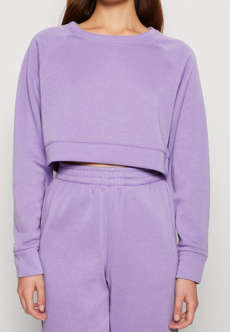 Femme portant un sweat court lavande assorti et un pantalon de survêtement taille haute avec taille élastique, debout devant un arrière-plan uni.