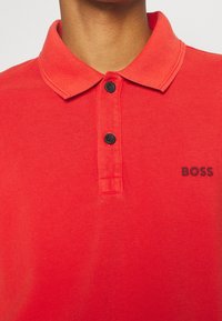 Röd polo skjorta med två svarta knappar och "BOSS"-logotyp på höger bröst, bärs av en person med medelbrun hud.