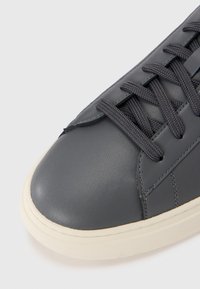 Grå lærersneaker med rund tå, teksturert overflate, kontrasterende lys såle og mørke flate snører. Har subtile sømdetaljer.