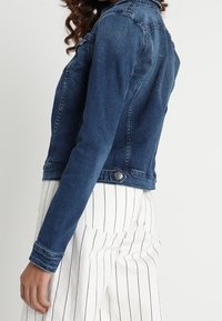 Tummansininen farkkutakki, jossa on vartalonmyötäinen muotoilu. Varustettu nappisuljennalla, rintataskuilla ja lyhennetty pituus. Yhdistetty raidallisiin housuihin.