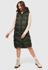 Grüne Stepp-Weste mit Kapuze, ausgestattet mit einem Reißverschluss und Druckknopfverschluss, knielang und mit durchgestepptem Muster. Getragen über einem elfenbeinfarbenen Pullover.