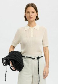 Polo shirt beige à côtes avec des manches courtes, présentant un col classique et trois boutons. Sac noir tissé tenu dans la main gauche. Pantalon léger bien ajusté.