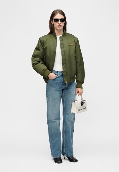 Άτομο που φοράει μαύρα γυαλιά ηλίου, πράσινο μπουφάν τύπου bomber, λευκό τοπ, μπλε τζιν, μαύρα παπούτσια, κρατώντας μια λευκή τσάντα χειρός με κείμενο και γραφικό.