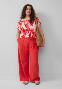 Eine Frau mit lockigem Haar trägt eine rot-weiß gemusterte Bluse, weite rote Hosen, Sandalen und trägt eine gewebte, beige Schultertasche.