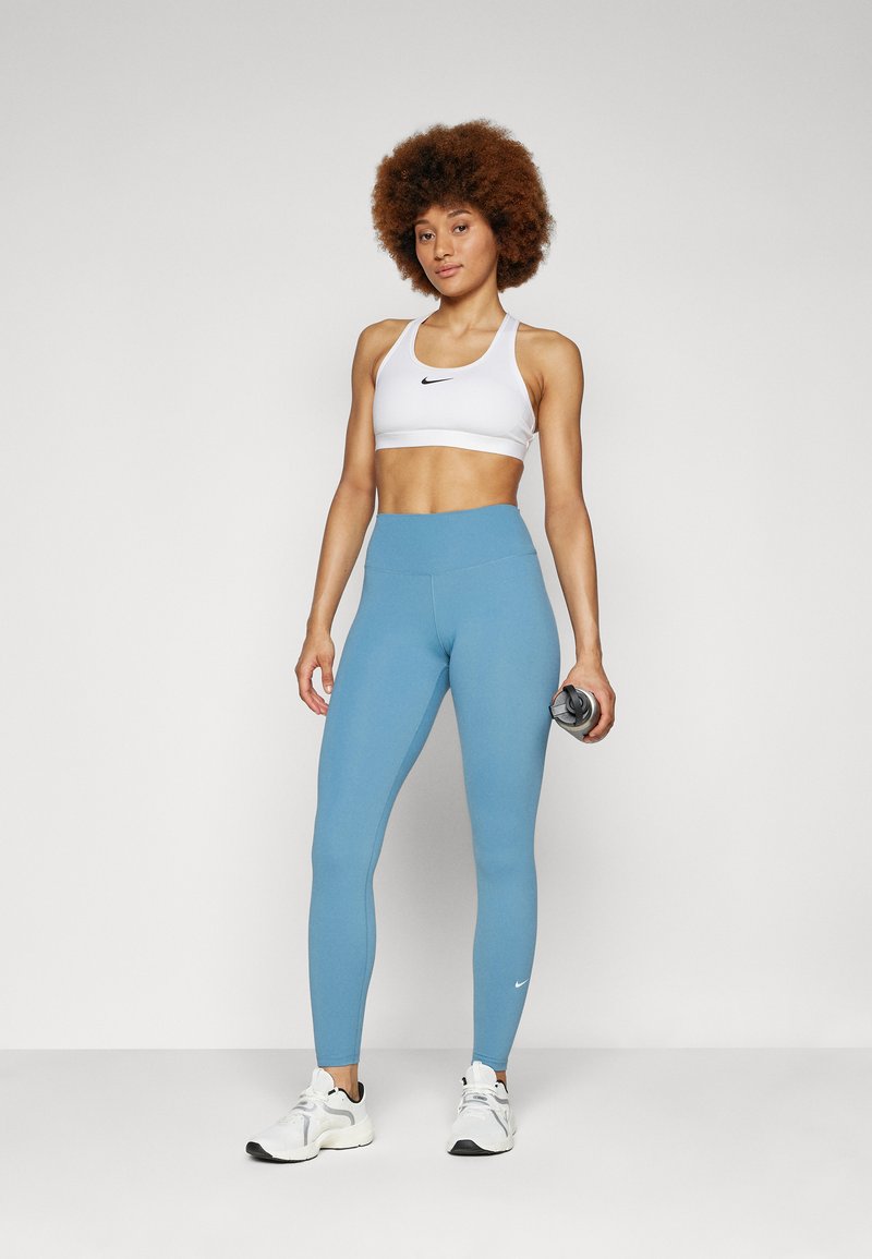 Soutien-gorge de sport blanc avec logo noir, associé à des leggings taille haute légers bleus. Le mannequin tient une bouteille d'eau, portant des chaussures de sport blanches.