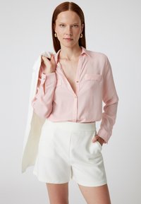 Hellrosa, knöpfbare Bluse mit zwei Brusttaschen, langen Ärmeln und lockerer Passform. Kombiniert mit weißen, hoch geschnittenen Shorts.