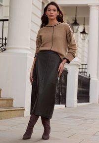 Béžový pletený svetr s žebrovanými manžetami a výstřihem, zkombinovaný s tmavě zelenou koženou midi sukní a fialovými semišovými vysokými botami na podpatku.