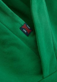 Gröna tyget med en mjuk textur, med en synlig etikett som har "Superdry" i rött på en svart bakgrund. Enkelt mönster, inga anmärkningsvärda mönster.