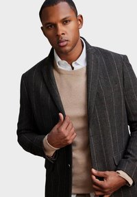Blazer a scacchi grigio scuro con righe bianche, con un solo bottone e due tasche frontali. Indossato sopra un maglione beige testurizzato.