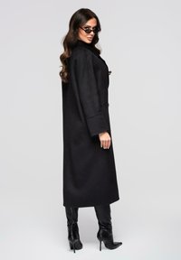 Manteau en laine noire présentant une coupe longue et droite, des manches surdimensionnées et un col haut, assorti à des bottes noires élégantes montant jusqu'aux genoux.
