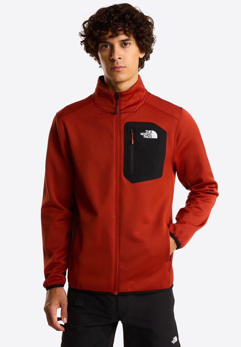 Rode softshell jas met een zwart ritsvak op de borst. Voorzien van hoge kraag en elastische manchetten. Gladde textuur, ontworpen voor buitengebruik.