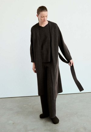 Mujer con suéter de punto marrón oscuro, pantalones anchos, cárdigan largo, sosteniendo un cinturón suelto en un entorno interior neutro.