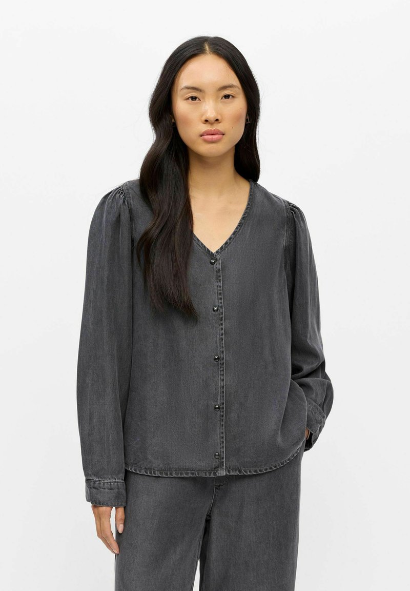 Jeune femme aux longs cheveux foncés portant un chemisier gris foncé boutonné et un pantalon assorti, debout devant un fond blanc uni.