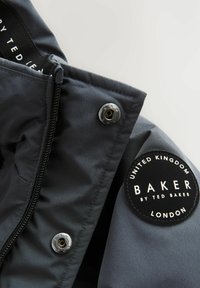 Veste grise avec fermeture éclair, pressions métalliques et un patch logo noir rond avec du texte blanc sur la manche.