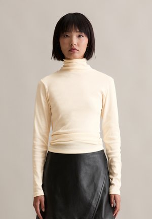 Cremfarbener Rollkragenpullover mit langen Ärmeln aus weichem, elastischem Stoff, kombiniert mit einem schwarzen Lederwickelrock mit subtiler Glanzoptik und seitlichen Details.
