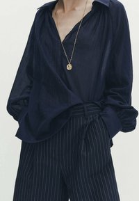 Osoba ubrana w luźną ciemnoniebieską koszulę wpuszczoną w ciemnoniebieskie spodnie w prążki, ozdobiona długim złotym naszyjnikiem z wisiorkiem.