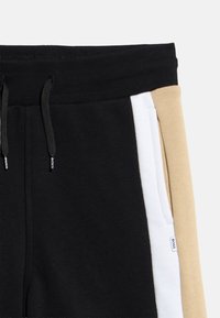 Κοντινό πλάνο μαύρων παντελονιών φόρμας με μαύρα κορδόνια, λευκή ρίγα στο πλάι, μπεζ πάνελ και μια μικρή ετικέτα της μάρκας BOSS δίπλα στην τσέπη.