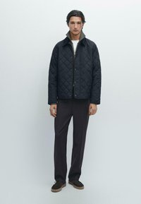 Quilted sort jakke med krave, med lynlås lukning, båret over en mørk skjorte og parret med mørke bukser og brune sko.