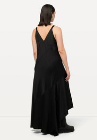 Robe noire sans manches avec un dos en V, présentant un ourlet asymétrique et une texture lisse. Portée avec des bottes noires jusqu'aux genoux.