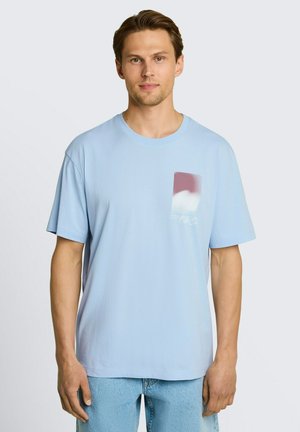 Mann mit hellbraunen Haaren, der ein lockeres hellblaues T-Shirt mit kleinem rechteckigem Wolkenmotiv auf der Brust und hellblaue Jeans trägt.