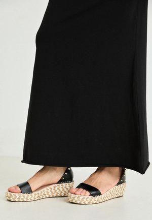 Pieds chaussés de sandales compensées noires avec des semelles en paille tressée et des brides noires à la cheville, associées à une longue jupe ou robe noire.