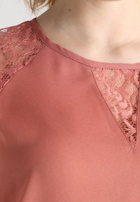 Różowa bluzka z koronkowymi akcentami na ramionach. Miękka, gładka tkanina z delikatnym połyskiem, z dekoltem w kształcie litery V i subtelnym koronkowym wykończeniem.