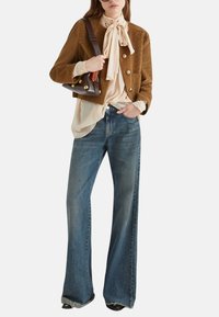 Giacca di velluto a coste marrone con bottoni dorati, blusa crema con fiocco, jeans svasati blu e scarpe nere. Borsa con dettagli in pelle scura.
