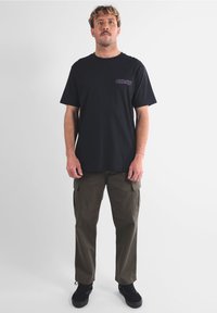 T-shirt nera in cotone con logo viola "OXBOW" sul lato sinistro del petto, abbinata a pantaloni cargo verde oliva e scarpe nere. Design minimalista.