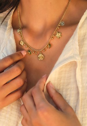 Gouden ketting met cirkelvormige en bloemen bedels, voorzien van kleine parels, een gestructureerde ketting en een gepolijste afwerking. Draag ik over een licht stoffen top.