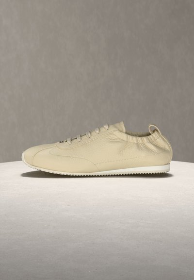 Μπεζ δερμάτινο καθημερινό αθλητικό παπούτσι με κορδόνια, ελαστικό γιακά και υφασμένη λευκή καουτσούκ σόλα σε στρογγυλή φωτεινή επιφάνεια.