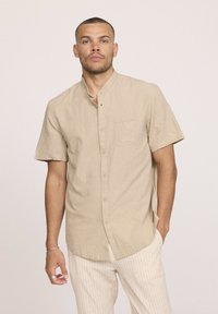 Chemise en lin beige à manches courtes avec un col montant, boutons sur le devant et une poche sur la poitrine gauche ; dotée d'une texture subtile et d'une coupe décontractée.