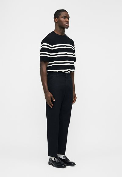 Άνδρας που στέκεται πλαγίως φορώντας μαύρο και άσπρο ριγέ κοντομάνικο πουκάμισο, μαύρο παντελόνι, λευκές κάλτσες και μαύρα παπούτσια φόρεμα.