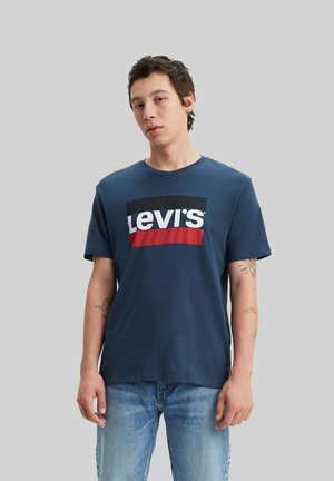Ung mand iført en marineblå Levi's t-shirt og lyseblå jeans, stående mod en ensfarvet grå baggrund.