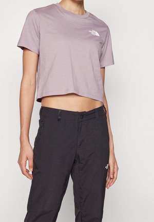 Person iført en cropped lavendelfarvet The North Face t-shirt og sorte bukser med lynlåslomme i siden og logo på låret.
