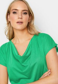 Top verde a maniche corte con scollo drappeggiato, texture morbida e fit rilassato. Il tessuto appare leggero e con una leggera lucentezza.