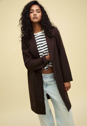 Long manteau marron en tissu doux, à l'avant ouvert, associé à une chemise rayée et un jean large bleu clair. Couleur unie sans matériel visible.