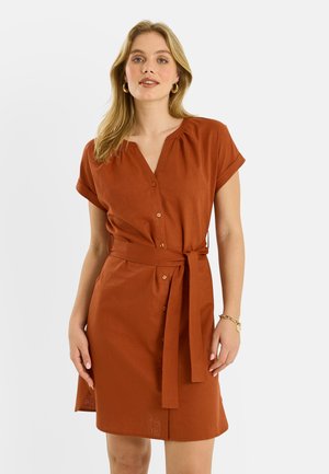 Mujer con vestido corto de botones y manga corta de color óxido con cinturón en la cintura, pendientes de aro dorados y una pulsera, de pie frente a un fondo liso.