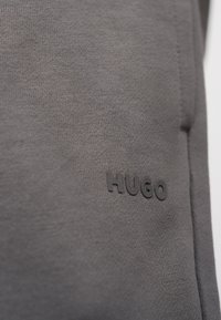 Κλείσιμο σε ύφασμα πλεκτό γκρι με μαύρο λογότυπο "HUGO" που είναι εγχάρακτο στην επιφάνεια, δείχνοντας υφή και λεπτομέρειες της ραφής του ενδύματος.