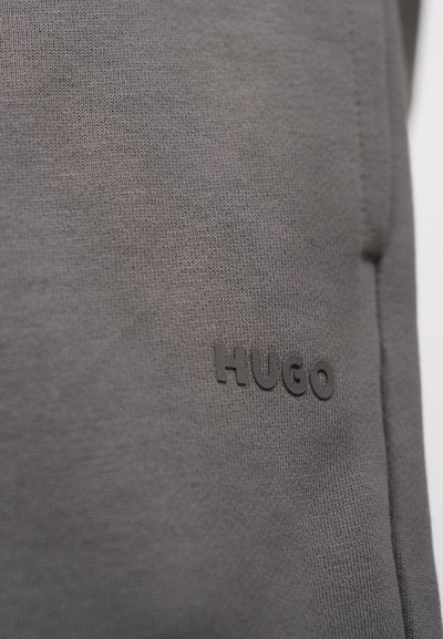 Gros plan sur un tissu gris tricoté avec le logo "HUGO" noir en relief sur la surface, montrant la texture et les détails des coutures du vêtement.