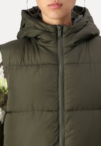 Puffervest i olivengrønn, med hette, frontglidelås og quiltet design. Stoffet ser glatt ut med en lett glans.
