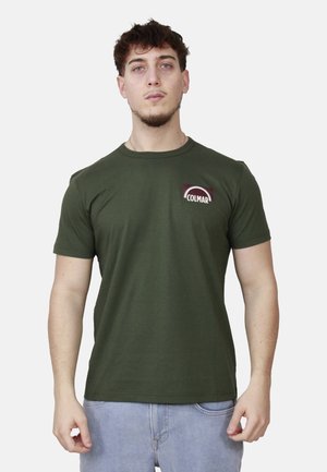 Giovane uomo che indossa una t-shirt Colmar verde oliva e jeans azzurri chiari, in piedi contro uno sfondo chiaro e uniforme.