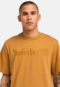 Bruine katoenen t-shirt met een geborduurd "Timberland"-logo en boomembleem. Korte mouwen en ronde halslijn. Gladde textuur.