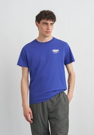 Jeune homme aux cheveux bouclés portant un t-shirt bleu Alpha Industries et un pantalon cargo gris, debout avec une main dans la poche devant un fond uni.