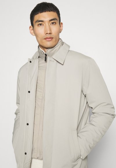 Chaqueta exterior gris claro con un cuello alto, botones visibles y cierre con cremallera oculta sobre una capa interior acolchada color crema. Tejido suave y resistente al agua.