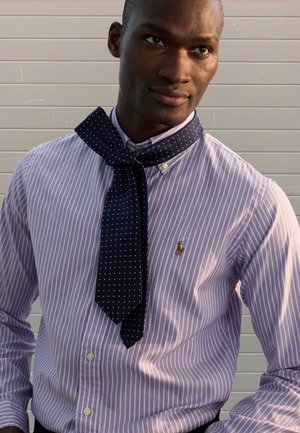 Camicia a righe viola chiaro con bottoni e piccolo logo. Una sciarpa a pois blu scuro annodata al collo aggiunge contrasto e dettaglio.