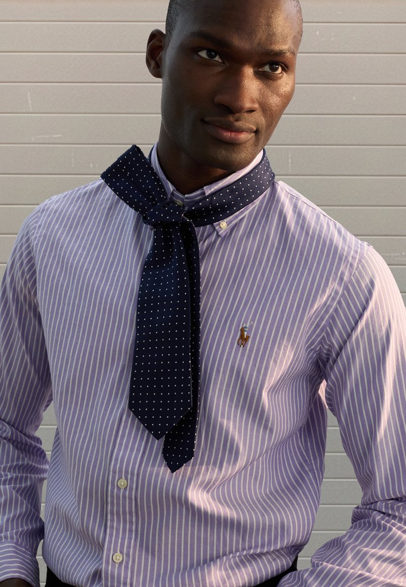 Šviesiai violetinė dryžuota palaidinė su sagomis ir mažo dydžio logotipu. Tamsiai mėlynos spalvos skarelė su baltais taškeliais, rišama prie kaklo, suteikia kontrastą ir detalumo.