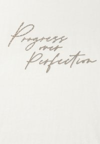 T-shirt blanc avec "Progrès plutôt que perfection" dans une police script argentée, présentant une texture lisse et une coupe légèrement ajustée.