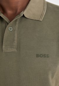 Olivi vihreä polo-paita, joka on valmistettu teksturoidusta puuvillasta, ja siinä on kaulus sekä kolme nappia. Rinnan kohdalla on "BOSS"-logo samassa värissä.