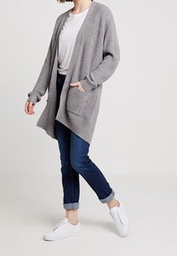 Szary dzianinowy kardigan z długimi rękawami, otwarty z przodu, z dwiema kieszeniami, zestawiony z ciemnymi dżinsami i białymi trampkami. Widoczny fakturowany wzór.